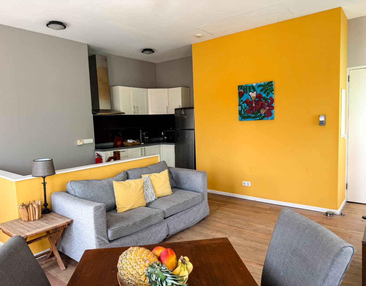 Open-plan living room with yellow accent wall, sofa, kitchen and dining table – Tierra apartment, Dushi Pietermaai